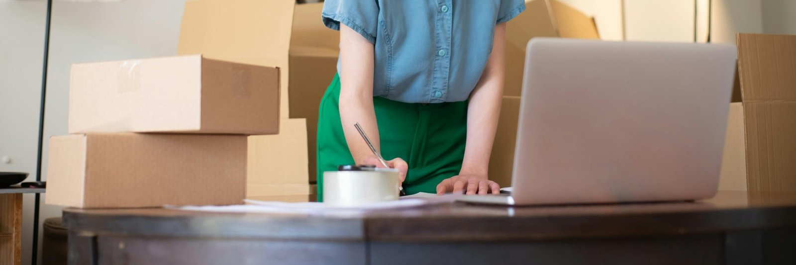 A woman working on her laptop surrounded by cardboard boxes, packing orders for he